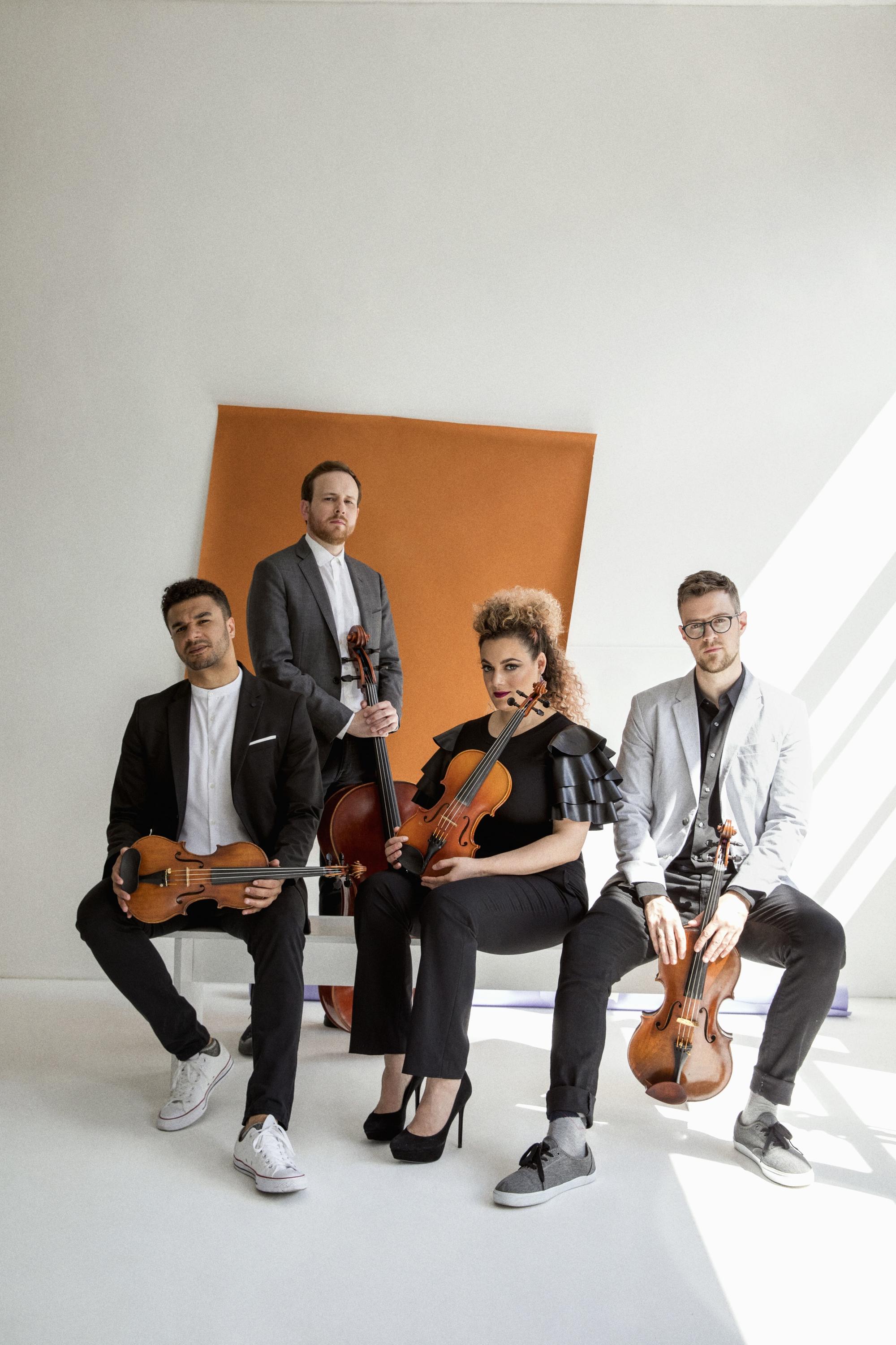Holding their instruments, three members of the PUBLIQuartet sit on a white bench in white room in front of an askew orange rectangle. The group's cellist stands behind them.