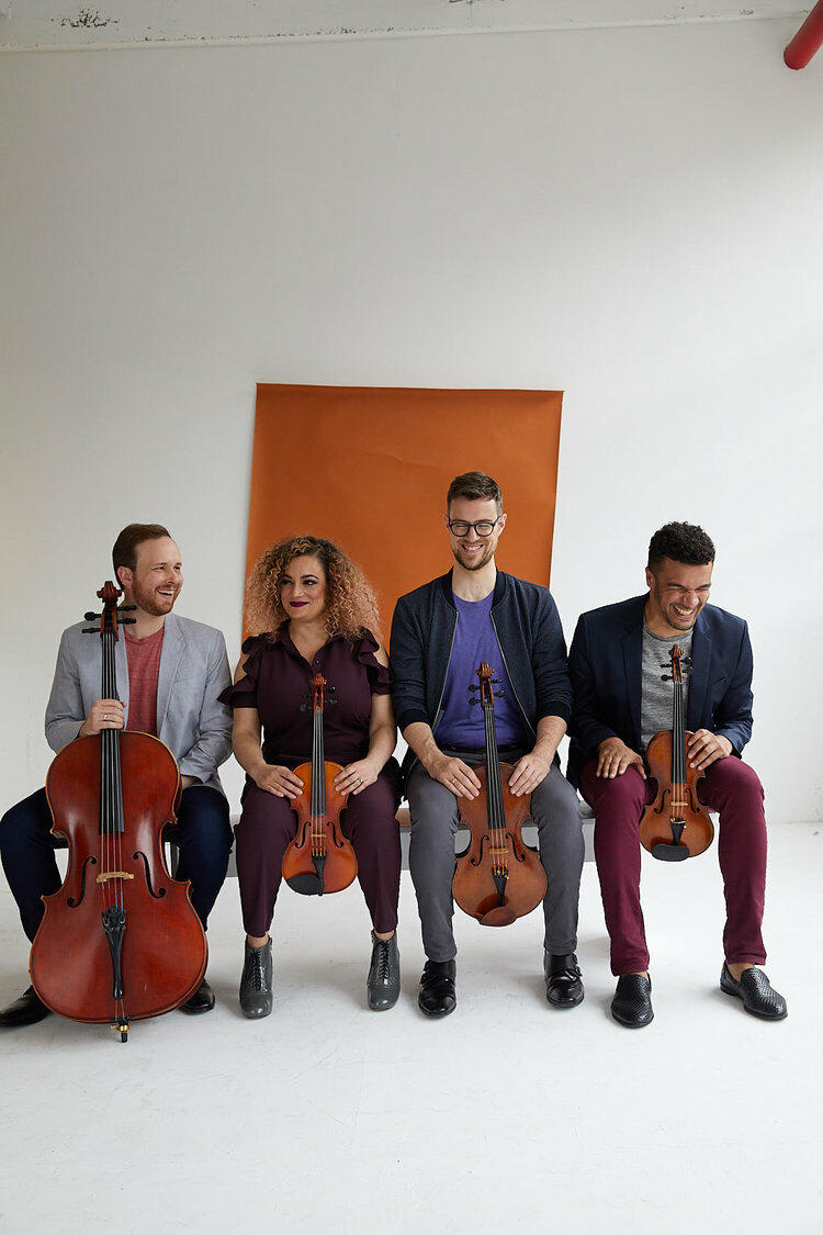 Image of a violin in the foreground and the smiling members of the Balourdet Quartet seated in the background