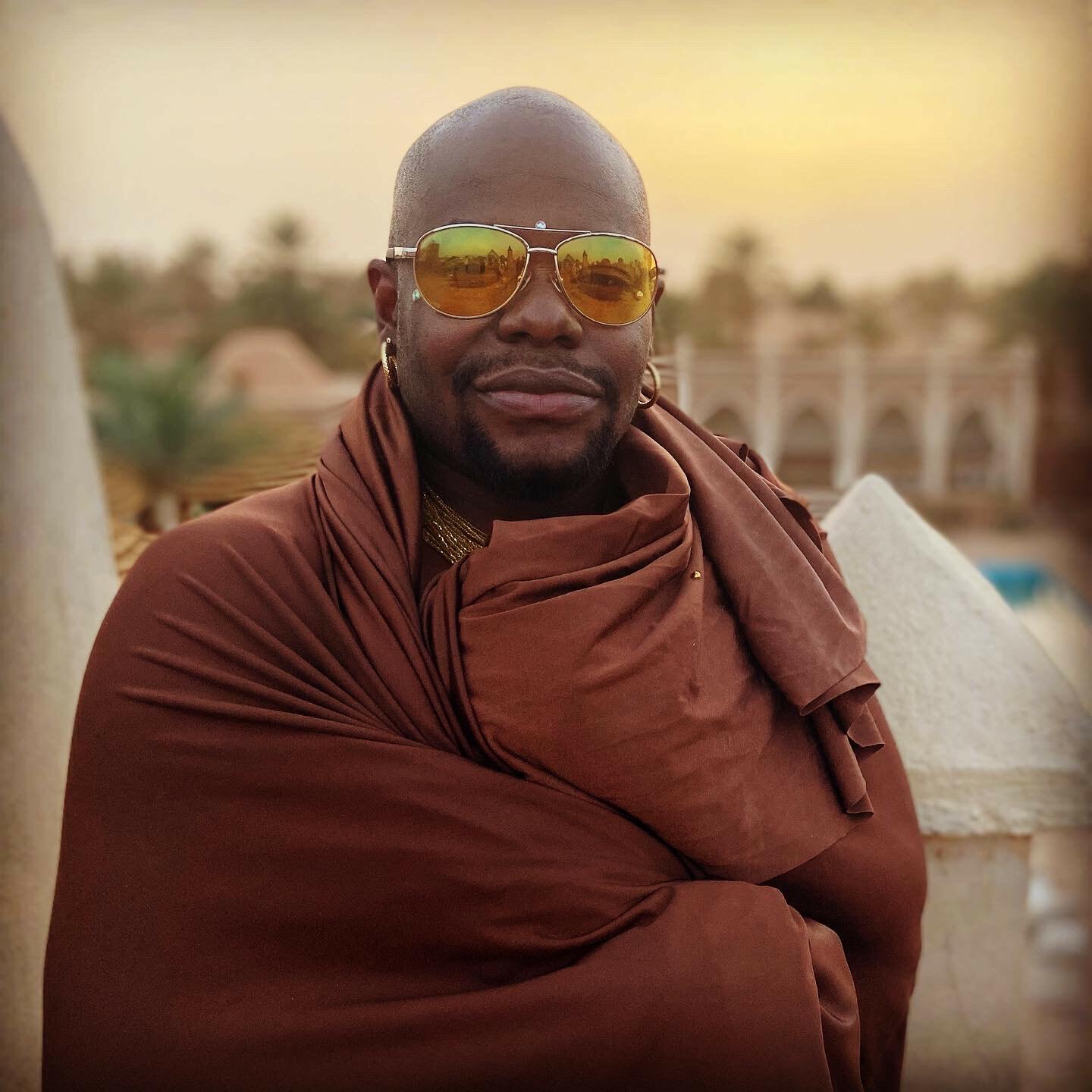 Jaamil is wearing yellow toned sunglasses and is wearing a brown robe which covers their body. The background consists of trees and architecture and the sun appears to be setting.