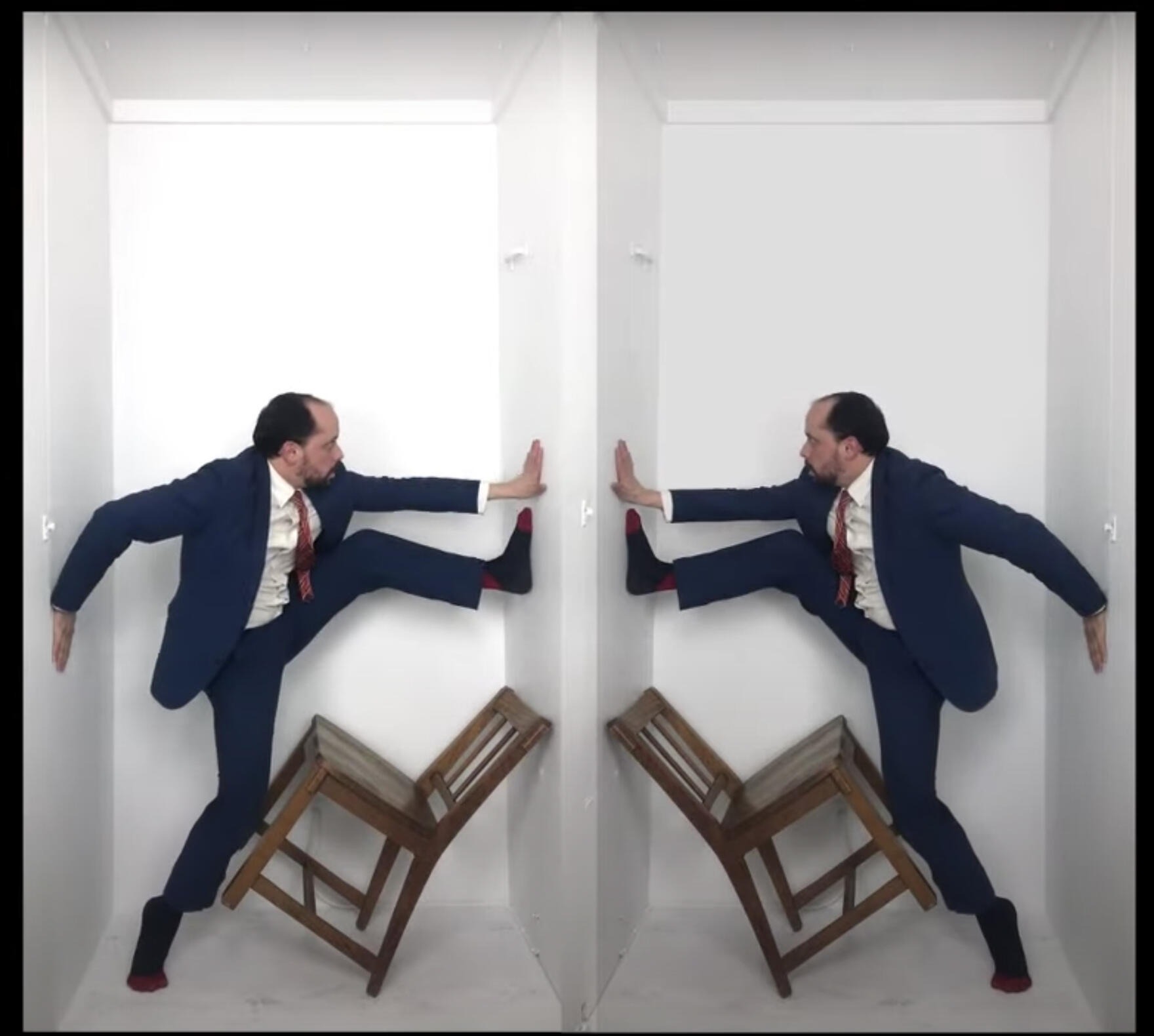 Mirrored images of Joshua William Gelb performing in his closet.  He a white man with dark hair and a beard, and is wearing a blue suit, white button-down shirt and reddish tie, and socks. His hands are on the walls of the closet, one foot on the floor and the other on one wall near his hand. His standing leg is pressed against a chair that is balancing on two legs against the wall. This image is constructed so that Gelb's foot, hand, and chair seem to be pressing against their mirror images. 