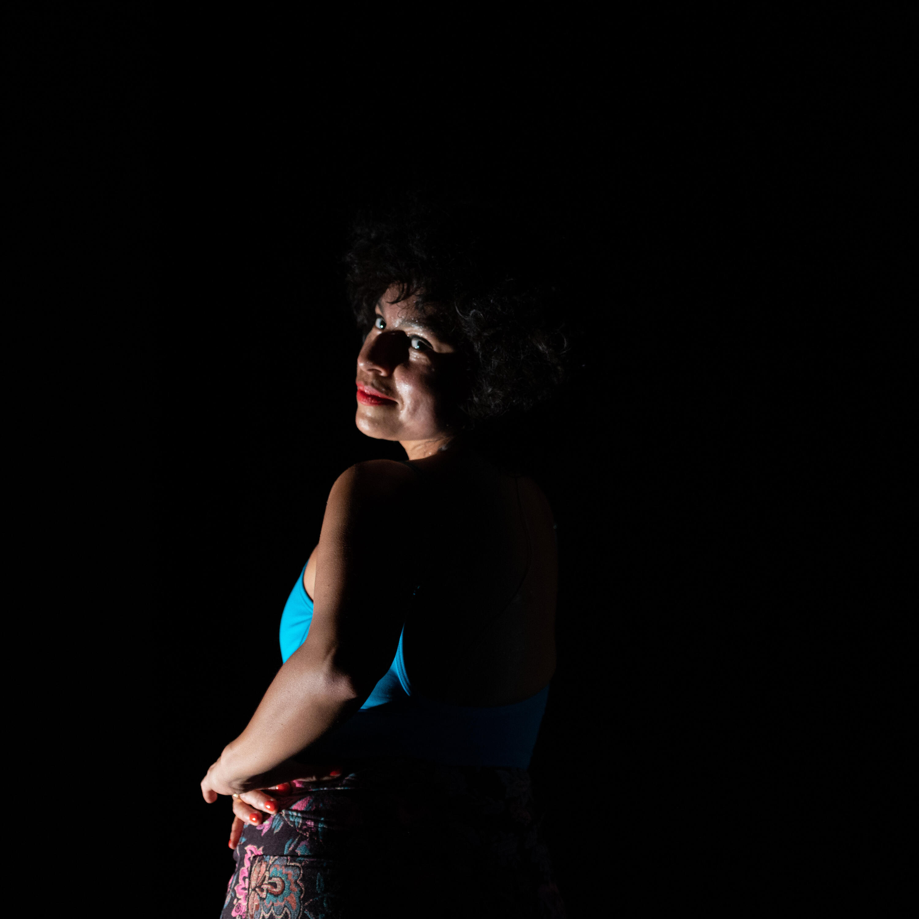  A woman with tan skin stands looking over her left shoulder with her left hand on her hip. She is smirking and has curly black hair in a bob with bangs. She wears a turquoise leotard and a pair of floral printed pants.