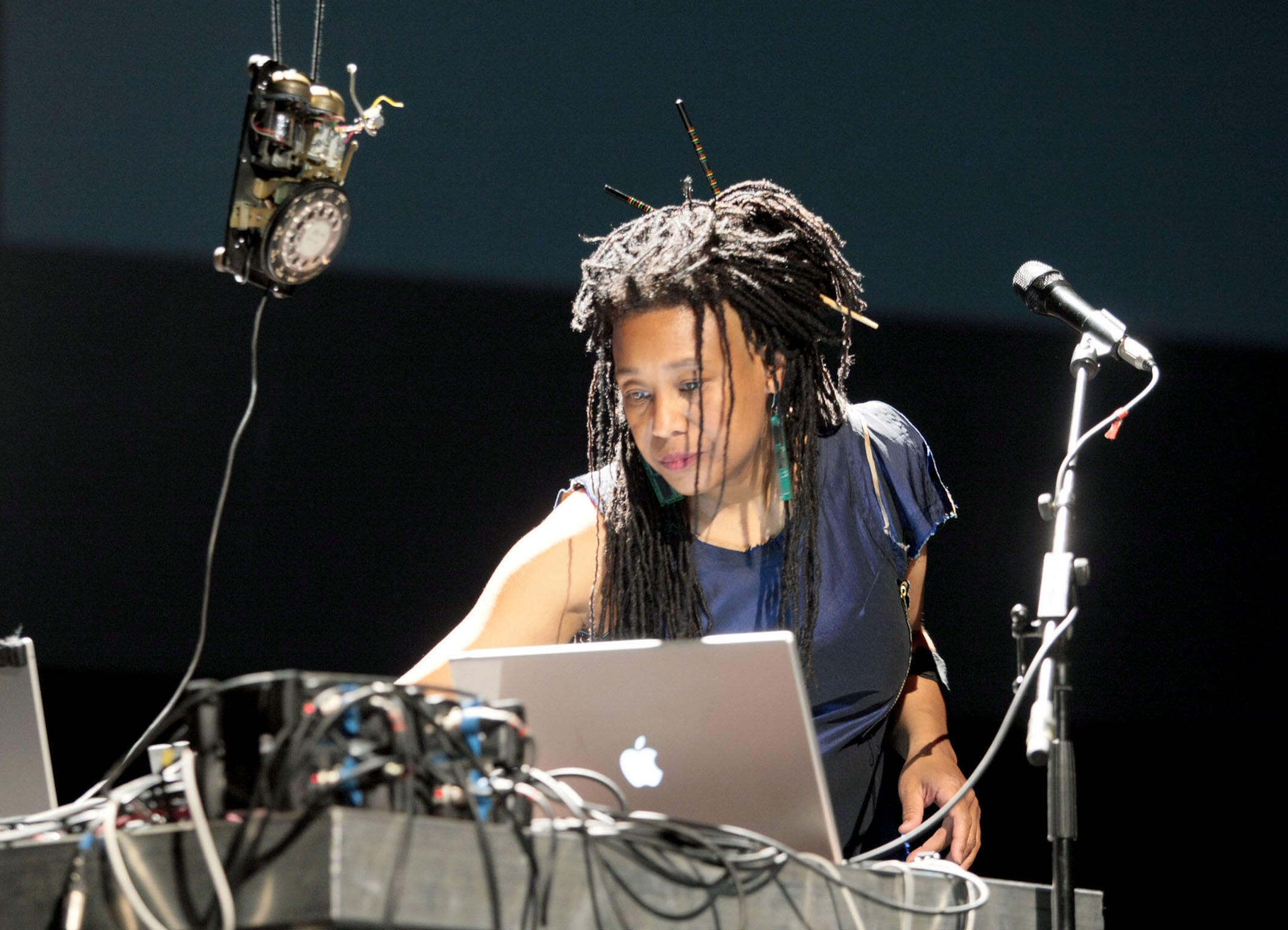 Pamela Z is on a stage surrounded by electronic equipment, microphones, and her laptop. Pamela wears her long hair in dreadlocks and is wearing an asymmetrical navy dress and turquoise earrings. 