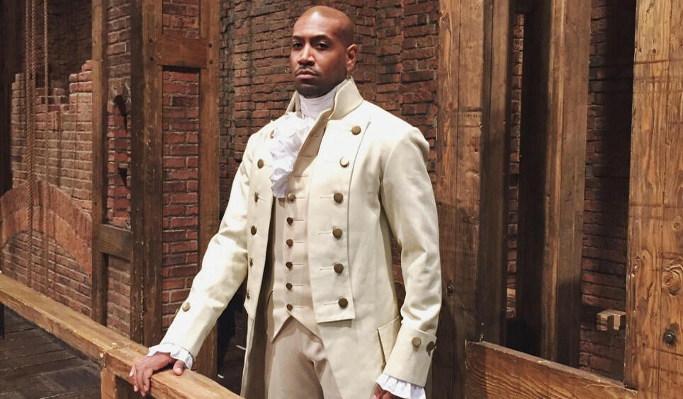 An image of Bryan Terrell Clark dressed in a period costume. He is standing somewhere on a theater stage among wooden beams. He gazes at the camera with a serious expression, one hand perched on a wooden ledge.