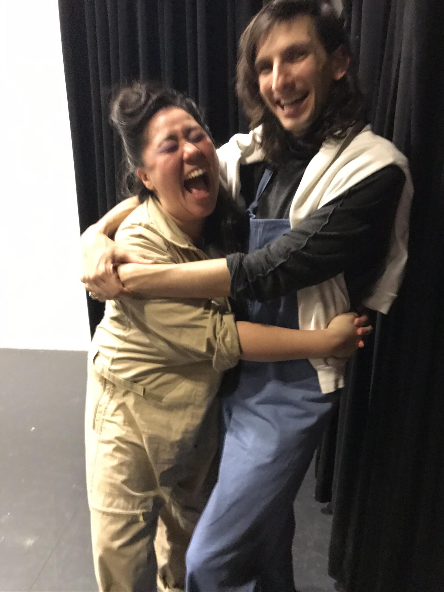 Image description: Una and Morgan embracing, their arms are holding each other, both are smiling laughing making the image blurry with joy. Morgan is wearing light blue overalls, a black long sleeve shirt, and a white sweatshirt draped over their shoulders, they are white and have dark brown wavy shoulder length hair. Una is wearing baggy khaki colored long sleeve overalls, they are Asian and have long black hair in a half up half down updo. There is a black curtain and black and white stage behind both of them.  