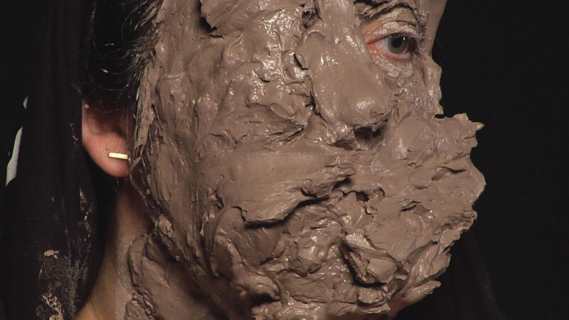 Emerging out of a black background is a cropped close-up of a performer's face, which is entirely covered in thick, gestural layers of mud or clay. One of the performer's eyes is exposed and peering out, while the other is completely consumed by the clay. You can see the edge of a black t-shirt, wrapped around the performer's hairline so that one of their ears is partially uncovered, revealing part of a gold earring. The rest of their face (mouth and nose included), are covered in expressive layers of mud.