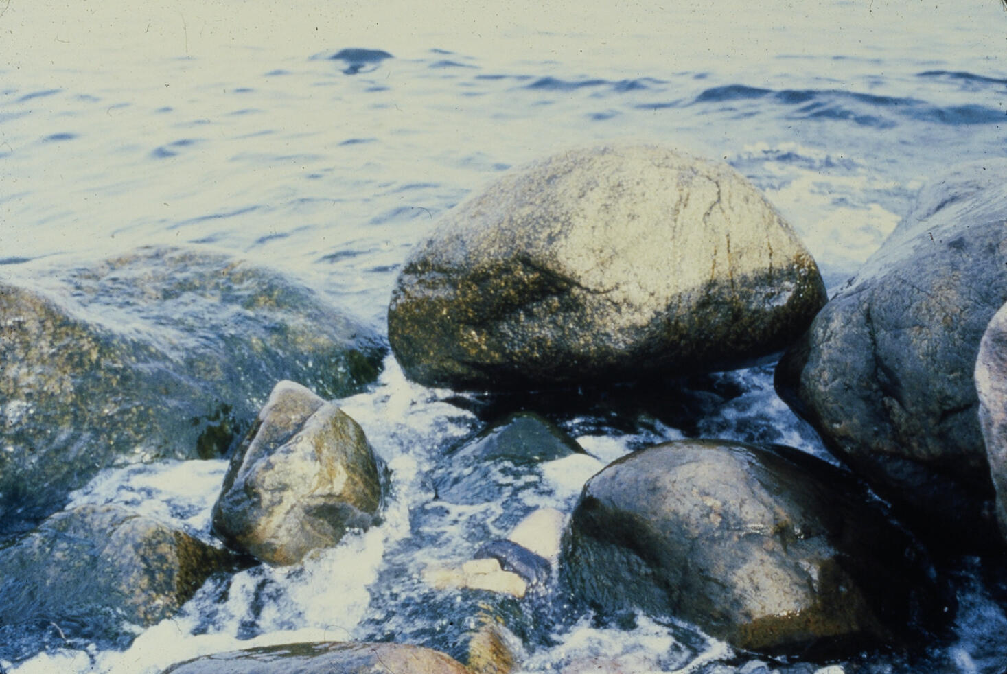 A color analog photograph showing a cluster of boulders and smaller stones at the seashore. Foamy sea water rushes between and across the stones, and where they are not fully submerged, they have a wet shine. Some of the smaller stones nestled at the center-front of the image have been left there as part of an artwork, but the arrangement is unstable, and it's not entirely clear where the artwork begins and ends.