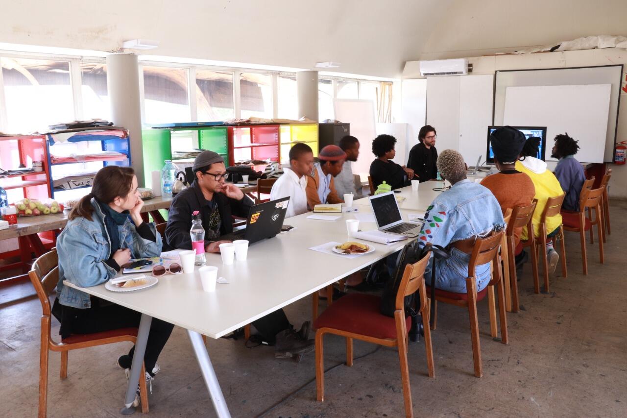 Image of eleven people sitting around a table, some with open computers and cups in front of them in white, airy room